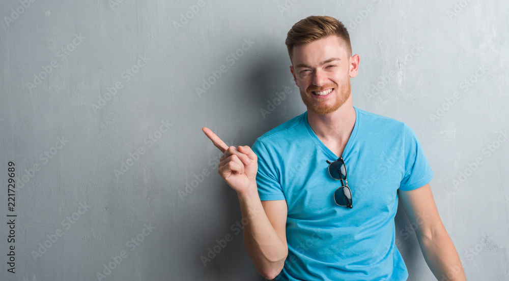 Young redhead man over grey grunge wall wearing casual outfit with a big smile on face, pointing with hand and finger to the side looking at the camera. Young redhead man over grey grunge wall wearing casual outfit with a big smile on face, pointing with hand and finger to the side looking at the camera.