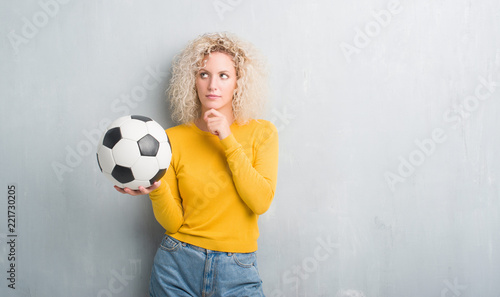 Young blonde woman over grunge grey background holding soccer football ball serious face thinking about question, very confused idea