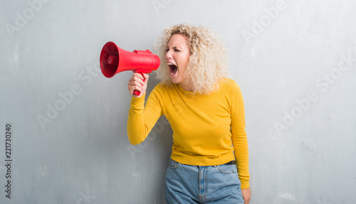 Young blonde woman over grunge grey background holding megaphone annoyed and frustrated shouting with anger, crazy and yelling with raised hand, anger concept