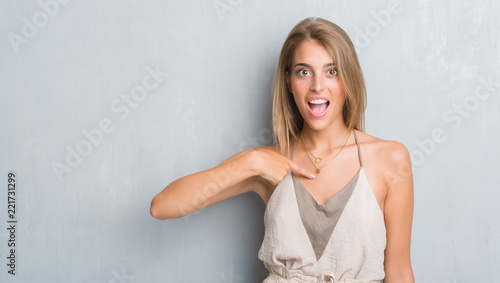 Beautiful young woman standing over grunge grey wall with surprise face pointing finger to himself