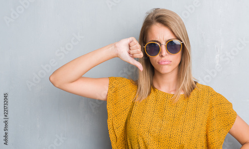 Beautiful young woman standing over grunge grey wall wearing retro sunglasses with angry face, negative sign showing dislike with thumbs down, rejection concept