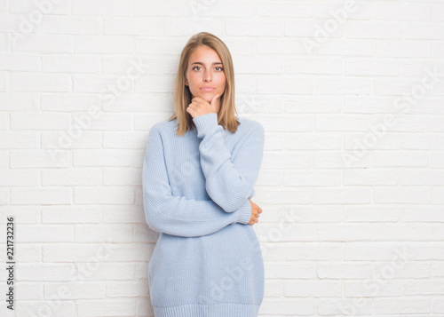 Beautiful young woman over white brick wall looking confident at the camera with smile with crossed arms and hand raised on chin. Thinking positive.
