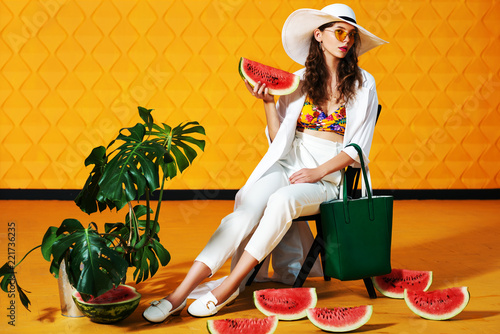 Full body studio fashion portrait of young beautiful model wearing long white trench coat, trousers, wide-brimmed hat, loafers, yellow sunglasses, holding green bag, posing on  background. Copy space