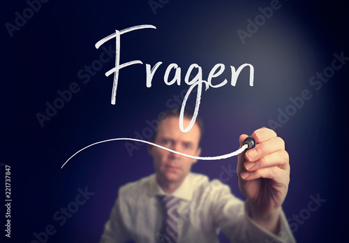 A businessman writing a Ask Questions "Fragen" concept in German with a white pen on a clear screen.