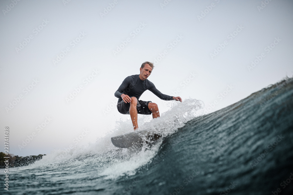 Young man wakesurfing on the board down the blue wave against the background of clear sky Young man wakesurfing on the board down the blue wave against the background of clear sky