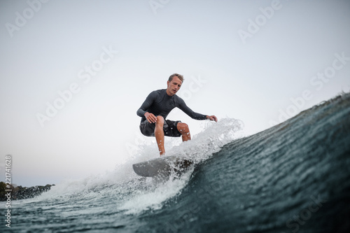 Young man wakesurfing on the board down the blue wave against the background of clear sky