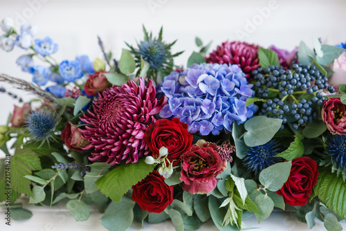Fototapeta Naklejka Na Ścianę i Meble -  Elongated floral arrangement in vintage metal vase. table setting. Blue and red color. Gorgeous bouquet of different flowers. Bright room, stucco on the wall