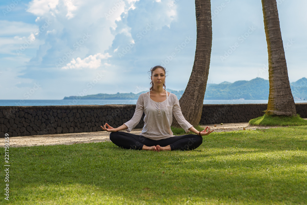 Beautiful young girl in leggings and tunic makes yoga practice, meditation on a beach of the ocean with green grass, yellow sand, azure water, blue sky, clouds. Sport healthy photo of model in Bali Beautiful young girl in leggings and tunic makes yoga practice, meditation on a beach of the ocean with green grass, yellow sand, azure water, blue sky, clouds. Sport healthy photo of model in Bali