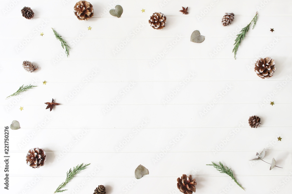 Christmas decorative composition. Floral frame of evergreen juniperus branches, eucalyptus leaves, pine, larch cones and glittering confetti stars. White wooden table background. Botanical pattern.