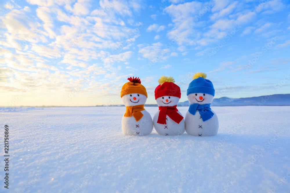 Fototapeta premium Three small snowman in colorful hats are standing on the snow. The scenery with mountains, field in snow, blue sky. Beautiful winter day.