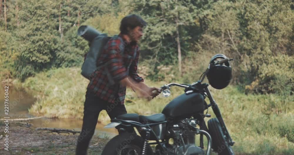 Hiker camping in forest. Guy biker with backpacker on his back sits on motorcycle in forest by river. Freedom lifestyle scene. 4K video shooting by handheld gimbal Hiker camping in forest. Guy biker with backpacker on his back sits on motorcycle in forest by river. Freedom lifestyle scene. 4K video shooting by handheld gimbal
