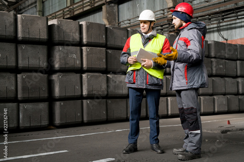 Full length  portrait of mature foreman wearing hardhat  giving instructions to worker while standing by concrete blocks in workshop, copy space photo