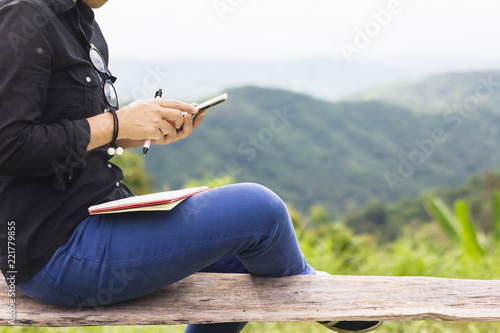 Asian girl siting and serfing the internet or playing game on mobile with green nature outdoor mountain background with diary book on her lap, concept of Internet communication photo