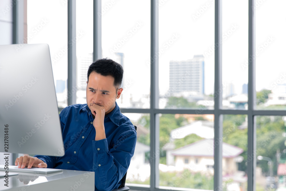 Young Asian businessman looking at the computer screen. His face expression was concerned with his work in modern office with large windows as background. Young Asian businessman looking at the computer screen. His face expression was concerned with his work in modern office with large windows as background.