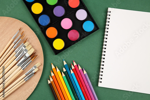 Top view of a set of watercolors, wooden palette, paint brushes, colorful pencils and blank sketchbook with copy space on a dark green background. Art, craft, school and education concept.