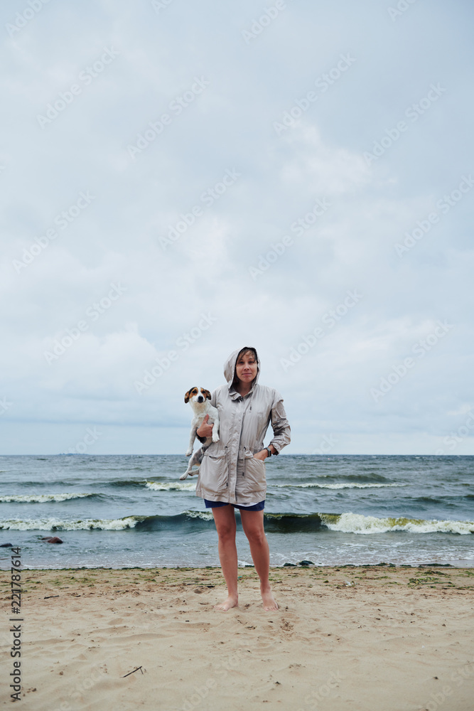Pretty young woman in warm jacket holding adorable dog and looking at camera while standing on sand near beautiful waving sea on cloudy day