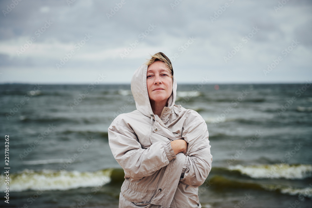 Pretty adult lady wrapping in warm jacket and looking at camera while standing on background of beautiful waving sea and overcast sky Pretty adult lady wrapping in warm jacket and looking at camera while standing on background of beautiful waving sea and overcast sky