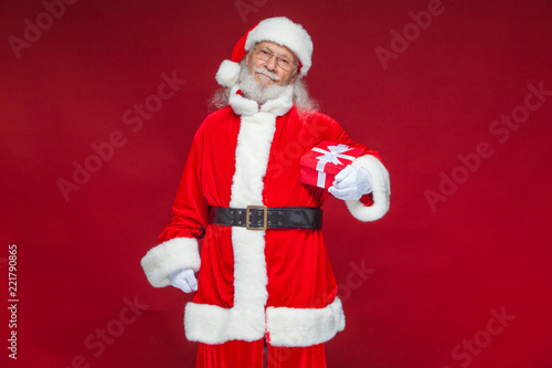 Christmas. Smiling Santa Claus in white gloves is holding a gift red box with a bow. Pointing at the gift. Isolated on red background.