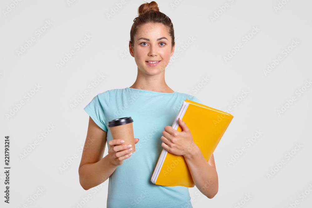 Shot of pleasant looking woman with hair bun has delighted expression, holds takeaway coffee and yellow folder, dressed casually, rests after having classes, isolated over white studio background Shot of pleasant looking woman with hair bun has delighted expression, holds takeaway coffee and yellow folder, dressed casually, rests after having classes, isolated over white studio background
