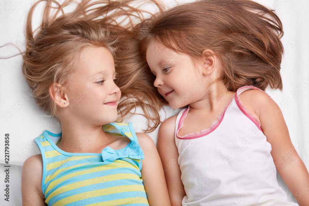 Two small sisters sleep together, look with positive expressions at each other, dressed in casual clothes, have good relationships. Best kid female friends have fun together in comfortable bed Two small sisters sleep together, look with positive expressions at each other, dressed in casual clothes, have good relationships. Best kid female friends have fun together in comfortable bed