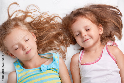 Top view of pleased attractive small girls or sisters smile together, sleep in bed, lie closely to each other, see pleasant dreams, dressed in nightwear. Children, relationship and friendship concept