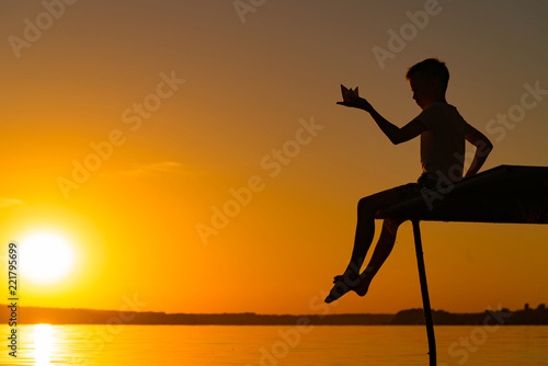 a little guy sits on a bridge and holds origami of paper ship in his hand in the evening at sunset. A boy plays with his toy near the river
