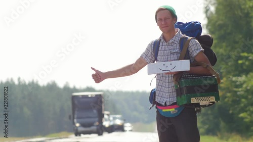 A man hitchhiker holding a white cardboard with a picture of a smile and honking passing cars