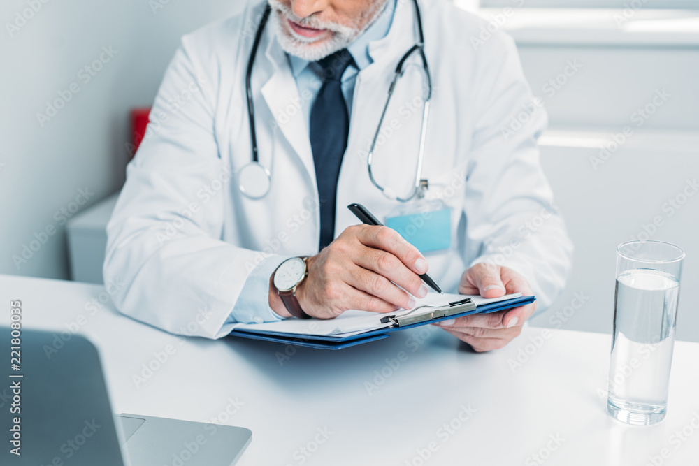 partial view of male doctor writing in clipboard and looking at laptop in office partial view of male doctor writing in clipboard and looking at laptop in office