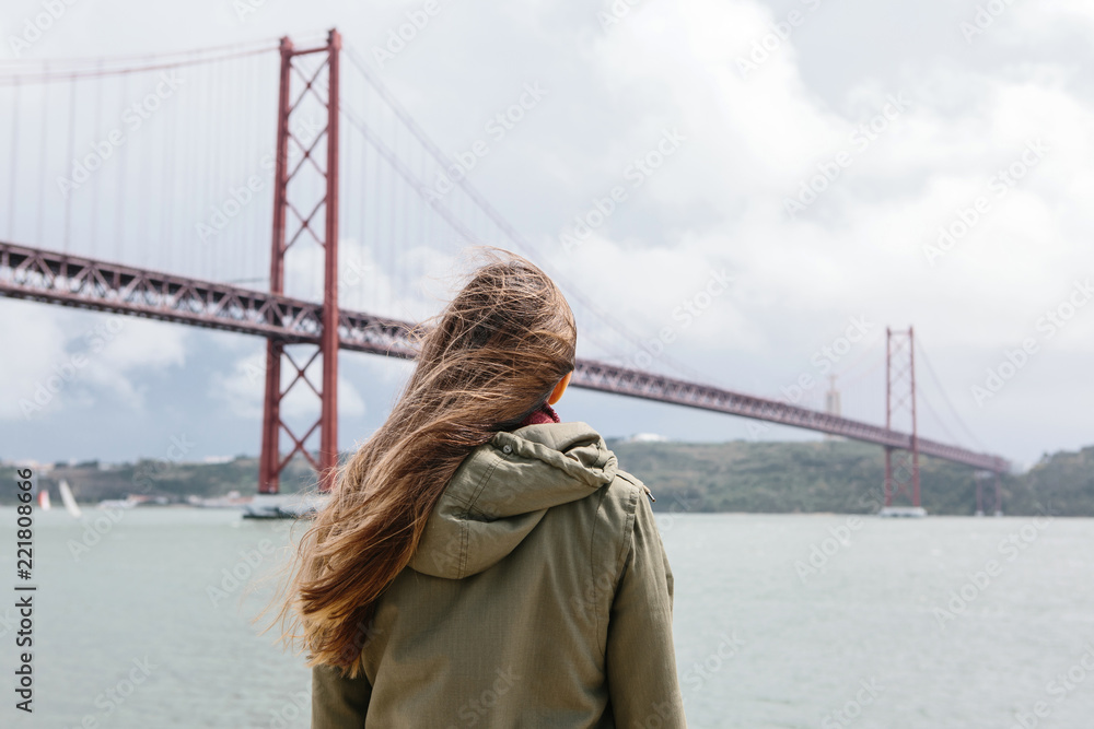 View from the back. The girl looks at the bridge called April 25 in Lisbon in Portugal in windy weather. View from the back. The girl looks at the bridge called April 25 in Lisbon in Portugal in windy weather.