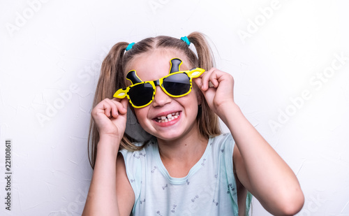 A smiling child girl in bright yellow sunglasses in the shape of a giraffe on a white background.