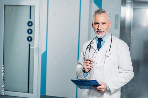smiling mature male doctor with stethoscope over neck holding eyeglasses with clipboard and looking at camera in hospital