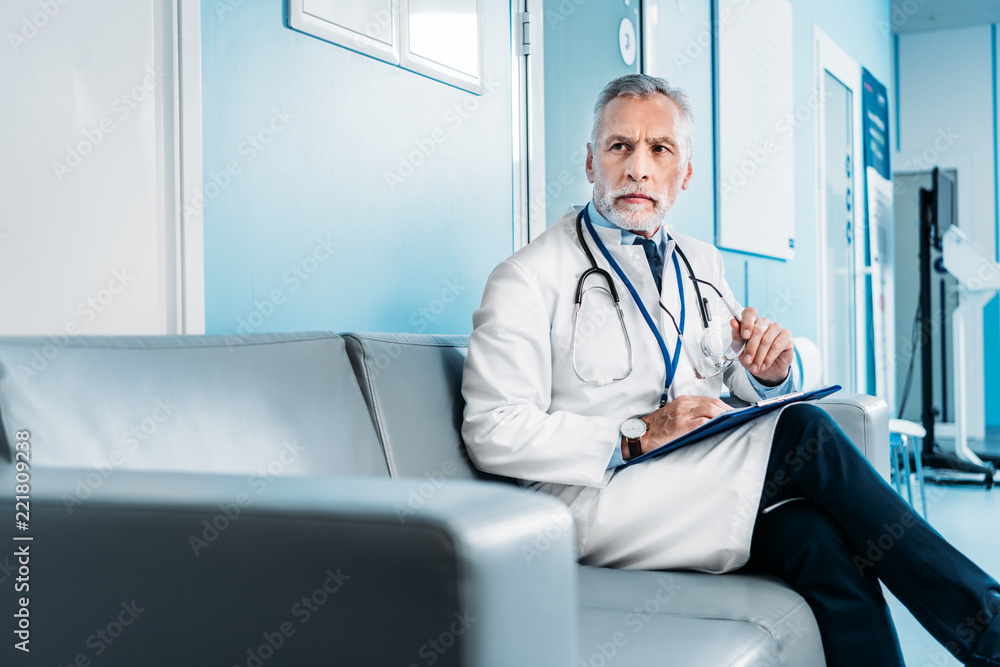middle aged male doctor with clipboard looking away and sitting on couch in hospital corridor