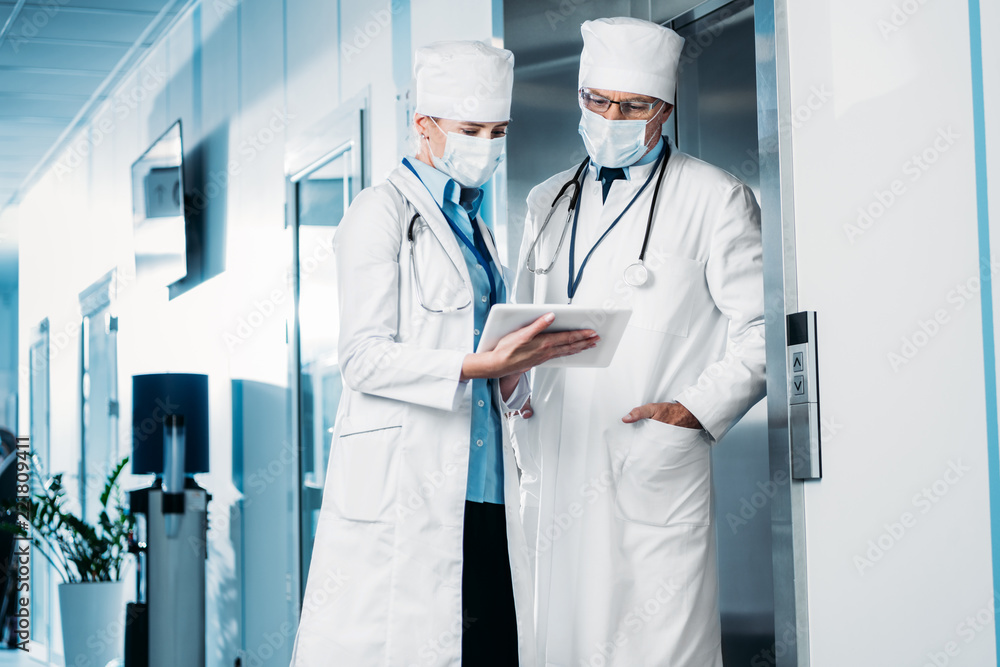 selective focus of female doctor in medical mask showing digital tablet to male colleague in hospital corridor selective focus of female doctor in medical mask showing digital tablet to male colleague in hospital corridor