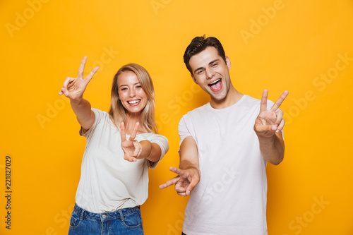 Excited young loving couple standing isolated over yellow wall background showing peace gesture.