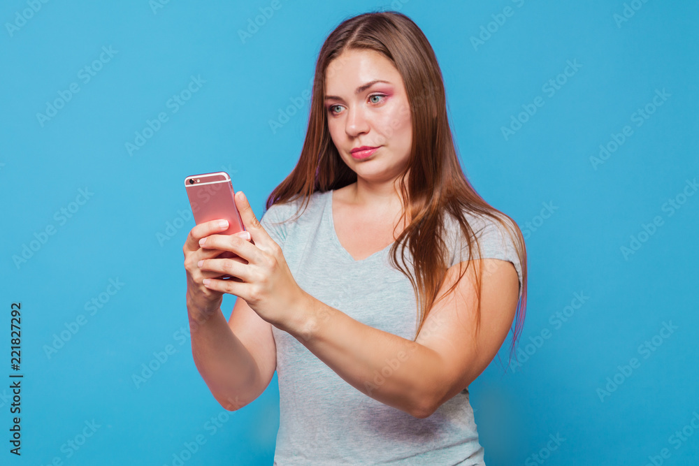 Young caucasian woman with brown hair and blue eyes looking to the pink phone with astonishment grimace and slight smile. Blue background, studio, isolated Young caucasian woman with brown hair and blue eyes looking to the pink phone with astonishment grimace and slight smile. Blue background, studio, isolated