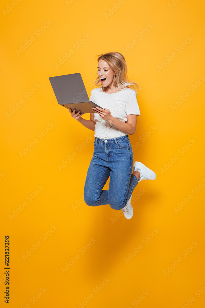 Excited young woman jumping isolated over yellow wall background using laptop computer. - obrazy, fototapety, plakaty 