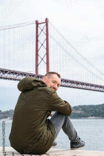 A young guy sits on the waterfront in Lisbon and looks at the camera