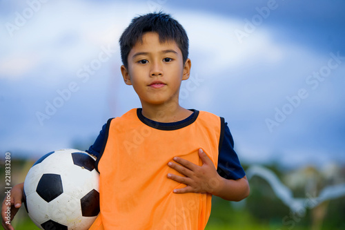 lifestyle portrait of handsome and happy young boy holding soccer ball playing football outdoors at green grass field smiling cheerful wearing training vest photo