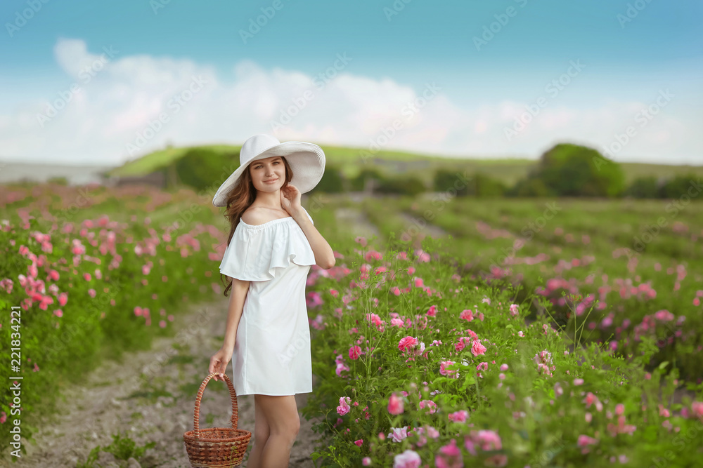 Beautiful young woman in white hat with long curly hair holding basket posing over spring blossom pink roses garden. Attractive charming brunette female outdoor portrait.