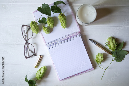 The opened notepad, pen, white candle, glasses and branches of hops as decoration on a white wooden table. Desktop still life with space for text.