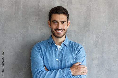 Horizontal shot of handsome Caucasian guy pictured isolated on grey background standing with arms crossed in casual clothes, looking confident and optimistic, smiling friendly and happily while posing