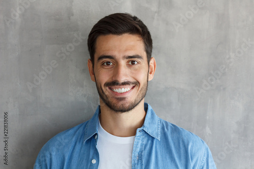 Horizontal headshot of young European male isolated on grey background wearing blue denim shirt in front of camera, smiling friendly and happily, satisfied with his life and free time activities