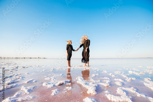 A stylish mother with her son in black clothes is walking among a pink salty lake.