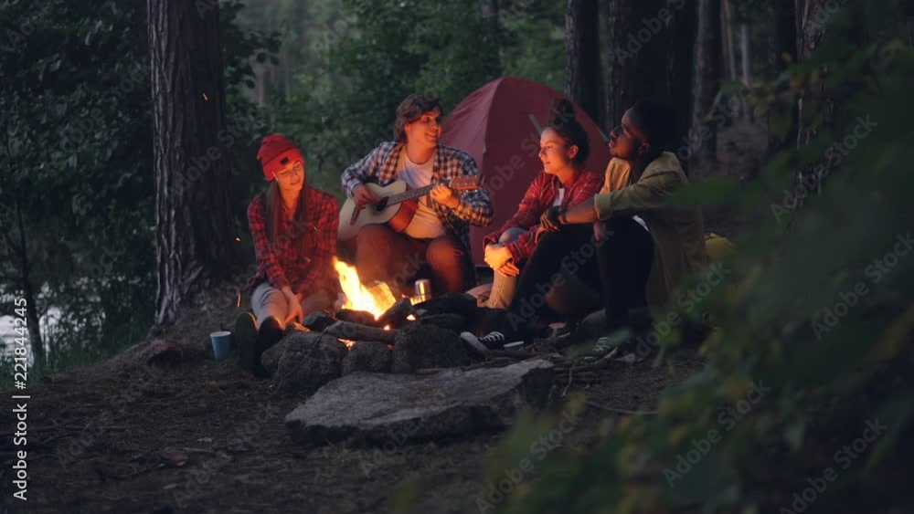 Girls and guys friends are singing songs while guitarist is playing musical instrument sitting near fire in forest and enjoying nature and music. Tent and lake are visible. Girls and guys friends are singing songs while guitarist is playing musical instrument sitting near fire in forest and enjoying nature and music. Tent and lake are visible.