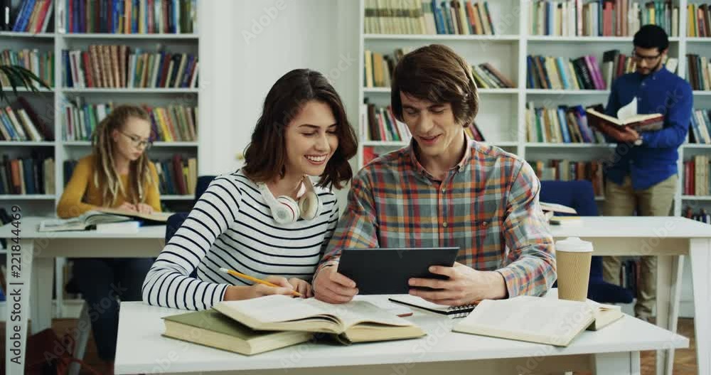 Caucasian couple of smiled students writing something and using tablet device while preparing for exams in the modern library. Caucasian couple of smiled students writing something and using tablet device while preparing for exams in the modern library.