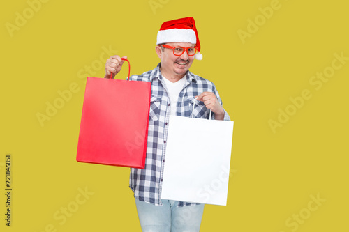 Happy modern middle aged man in casual style and red new year santa cap, eyeglasses and shirt, holding shopping packages with toothy smile, looking at camera. indoor, isolated on yellow background