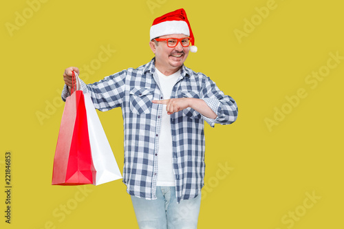 Casual styled funny middle aged man in red new year santa cap, eyeglasses standing and pointing finger to shopping bags and toothy smile, looking at camera. Studio shot, isolated on yellow background