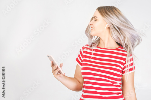 Happy young woman in earphones is listening to music with smart phone, dancing, having fun and smiling. White background, isolated.