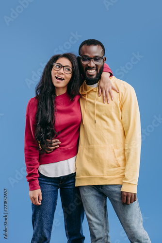 stylish happy african american couple in eyeglasses embracing each other isolated on blue background