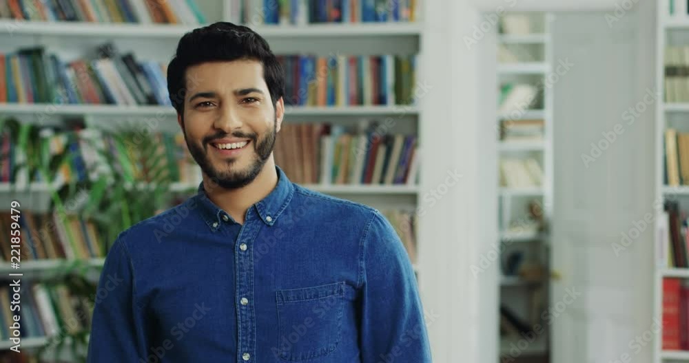 Portrait shot of the Arabian handsome young man standing in the modern library, looking at the side and turning head to the camera with a smile. Portrait shot of the Arabian handsome young man standing in the modern library, looking at the side and turning head to the camera with a smile.